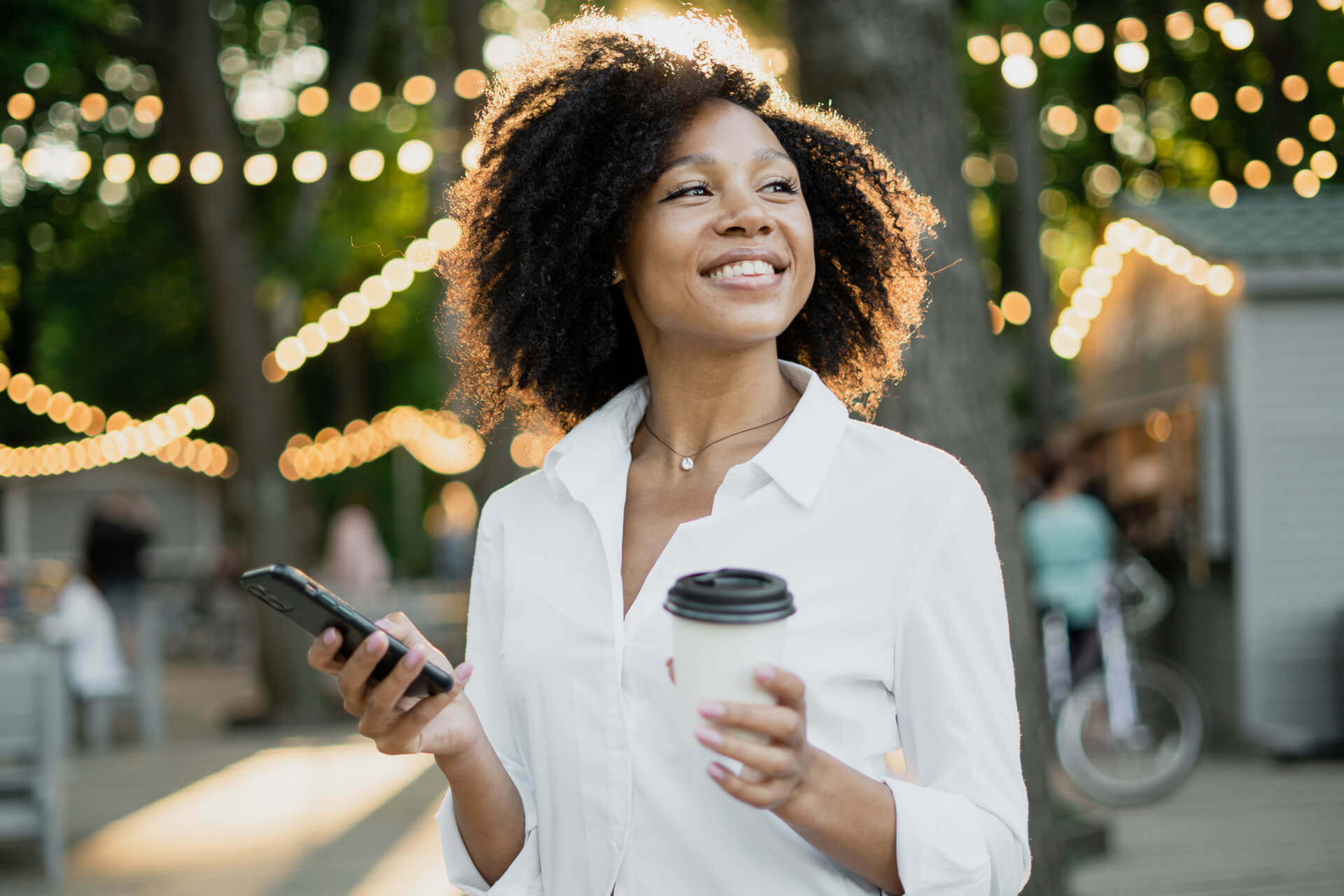 Black woman in white shirt holding phone and coffee in outside space with string lights 